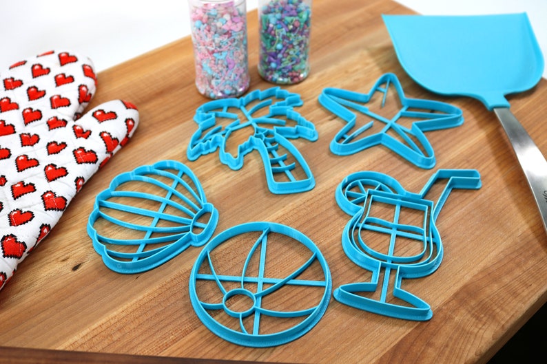 May include: A set of blue plastic cookie cutters shaped like a palm tree, seashell, starfish, beach ball, and cocktail glass. The cutters are on a wooden cutting board with two jars of sprinkles in the background.