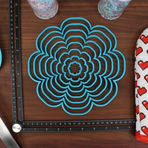 May include: A set of seven blue plastic flower-shaped cookie cutters of varying sizes. The cutters are arranged in a circle on a wooden surface.