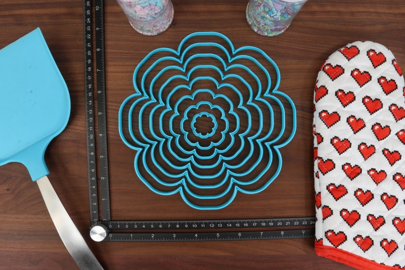 May include: A set of seven blue plastic flower-shaped cookie cutters of varying sizes. The cutters are arranged in a circle on a wooden surface.