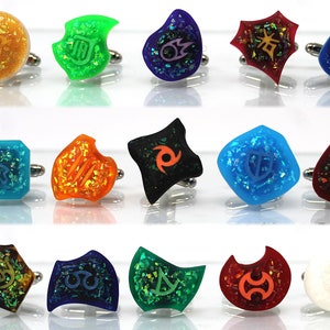 May include: Set of 14 colorful cufflinks with various symbols and designs. Each cufflink features a different symbol, including a shield, a crescent moon, a star, and a swirl. The cufflinks are made of resin and have a glittery finish.