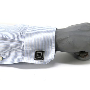 Puede incluir: Una camisa blanca de vestir con rayas azules y un gemelo plateado con un s&iacute;mbolo en blanco y negro.