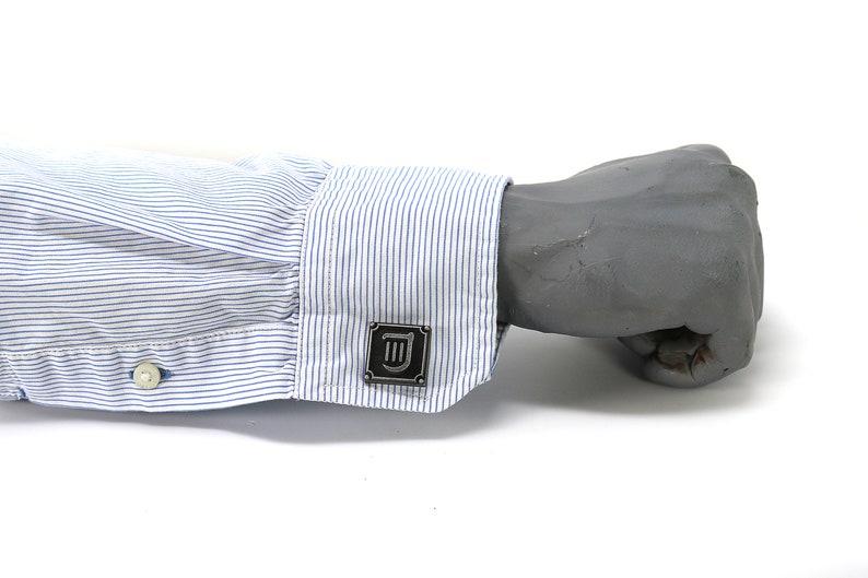 Puede incluir: Una camisa blanca de vestir con rayas azules y un gemelo plateado con un s&iacute;mbolo en blanco y negro.