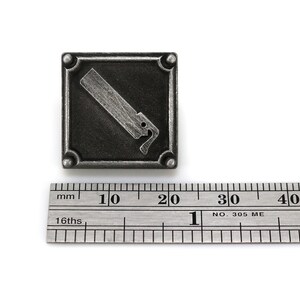 May include: A silver-toned metal square pin with a raised image of a saw. The pin is approximately 2 cm wide.