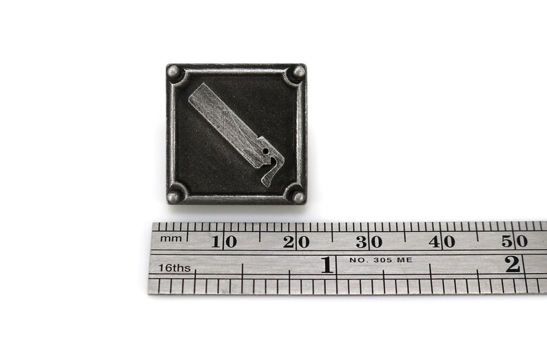 May include: A silver-toned metal square pin with a raised image of a saw. The pin is approximately 2 cm wide.
