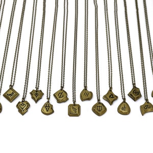 Pode incluir: Um conjunto de 16 colares dourados com diferentes designs de pingentes. Cada pingente apresenta um s&iacute;mbolo ou design &uacute;nico, incluindo um escudo, uma lua crescente, uma estrela e um c&iacute;rculo. Os colares est&atilde;o todos sobre um fundo branco.
