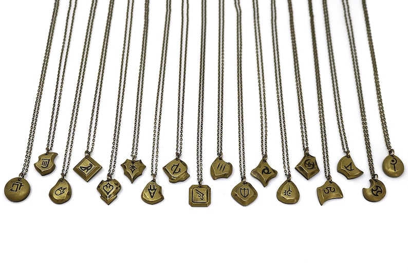 Pode incluir: Um conjunto de 16 colares dourados com diferentes designs de pingentes. Cada pingente apresenta um s&iacute;mbolo ou design &uacute;nico, incluindo um escudo, uma lua crescente, uma estrela e um c&iacute;rculo. Os colares est&atilde;o todos sobre um fundo branco.