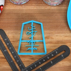 May include: A light blue plastic cookie cutter in the shape of a Japanese character, with the character's name in the center. Two black rulers and a blue spatula are also visible. A white and red heart-patterned oven mitt is in the background.