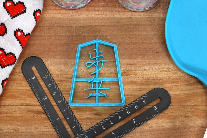 May include: A light blue plastic cookie cutter in the shape of a Japanese character, with the character's name in the center. Two black rulers and a blue spatula are also visible. A white and red heart-patterned oven mitt is in the background.