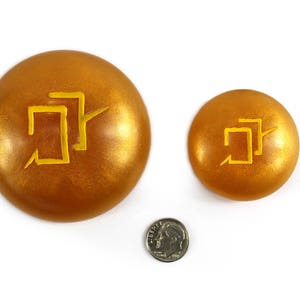 Puede incluir: Dos discos decorativos redondos de color dorado con un s&iacute;mbolo amarillo en cada uno. El s&iacute;mbolo es un dise&ntilde;o geom&eacute;trico estilizado.