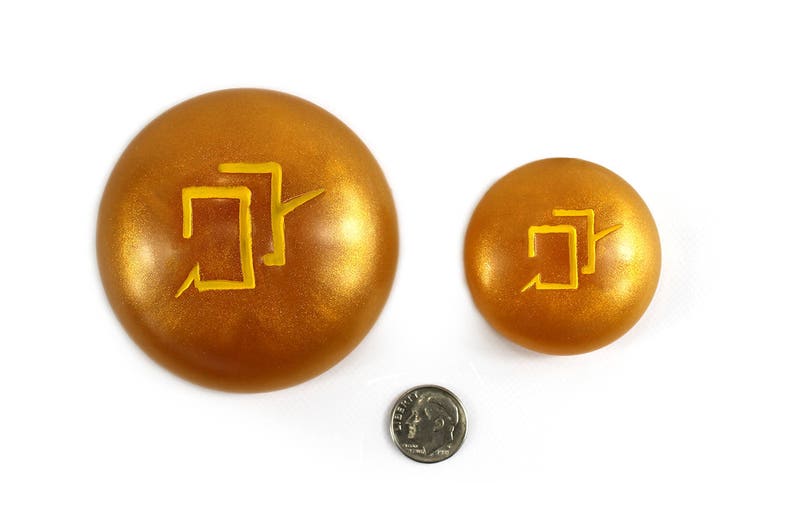 Puede incluir: Dos discos decorativos redondos de color dorado con un s&iacute;mbolo amarillo en cada uno. El s&iacute;mbolo es un dise&ntilde;o geom&eacute;trico estilizado.