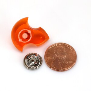 May include: Orange plastic pinback button shaped like a crescent moon with a silver pin on the back. The button is approximately the size of a UK penny.