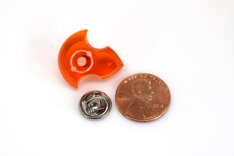 May include: Orange plastic pinback button shaped like a crescent moon with a silver pin on the back. The button is approximately the size of a UK penny.