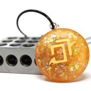 May include: A gold pendant necklace with a yellow and orange resin charm featuring a white geometric symbol. The charm is set against a background of iridescent glitter.
