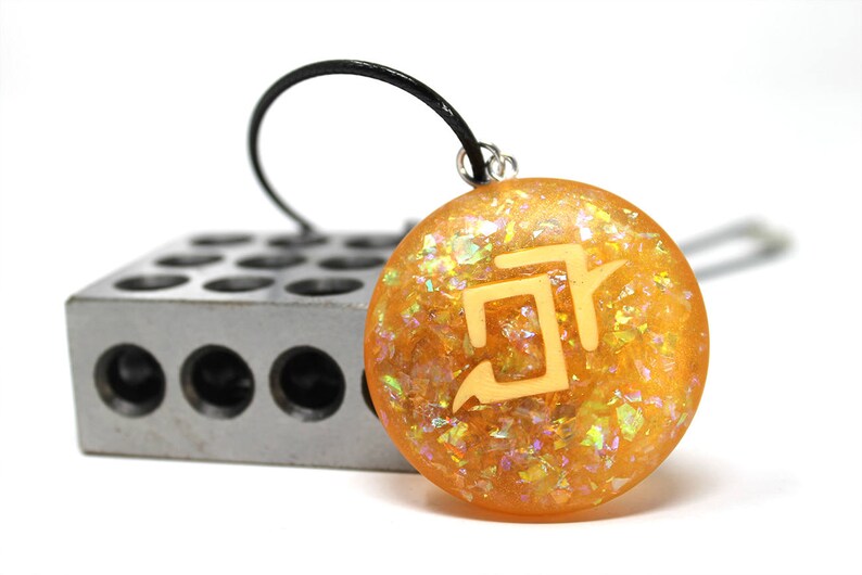 May include: A gold pendant necklace with a yellow and orange resin charm featuring a white geometric symbol. The charm is set against a background of iridescent glitter.