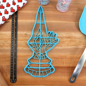 May include: Blue plastic cookie cutter shaped like a hand in a prayer position. The cutter is on a wooden surface with a ruler, a rolling pin, and a blue spatula.