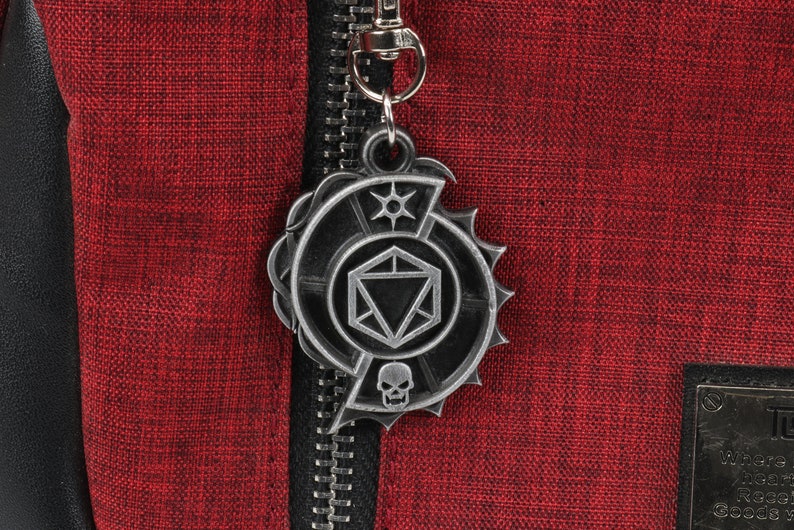 May include: A silver metal zipper pull with a Dungeons and Dragons themed design. The design features a 20-sided die, a skull, and a star.