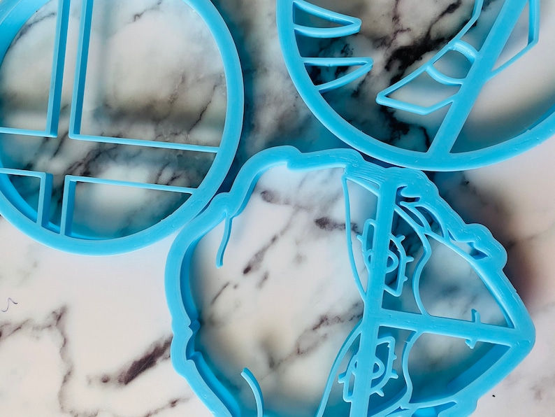 May include: Three blue plastic cookie cutters, one shaped like a circle with a grid pattern, one shaped like a flower with a swirl pattern, and one shaped like a geometric design.