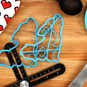 May include: Two blue plastic cookie cutters shaped like a wolf howling at the moon. The cutters are on a wooden surface with a ruler and a blue spatula.