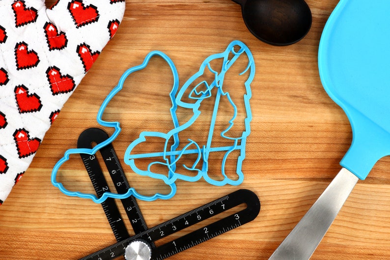 May include: Two blue plastic cookie cutters shaped like a wolf howling at the moon. The cutters are on a wooden surface with a ruler and a blue spatula.