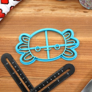 May include: A blue plastic cookie cutter shaped like an axolotl with a smiling face. The cutter has a cross-shaped design inside the axolotl's body.