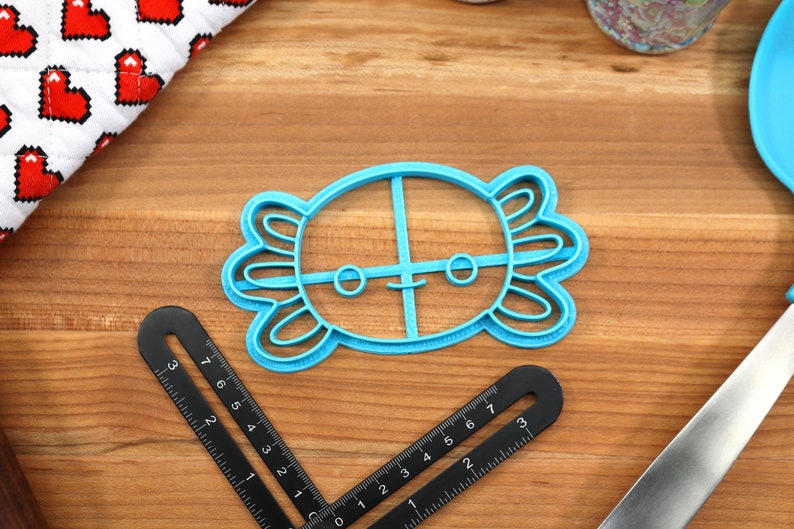 May include: A blue plastic cookie cutter shaped like an axolotl with a smiling face. The cutter has a cross-shaped design inside the axolotl's body.