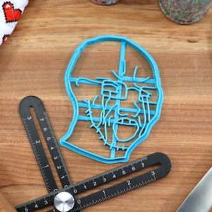 May include: A blue plastic cookie cutter shaped like a face with a detailed design. The cutter is on a wooden surface with a ruler and a spatula.