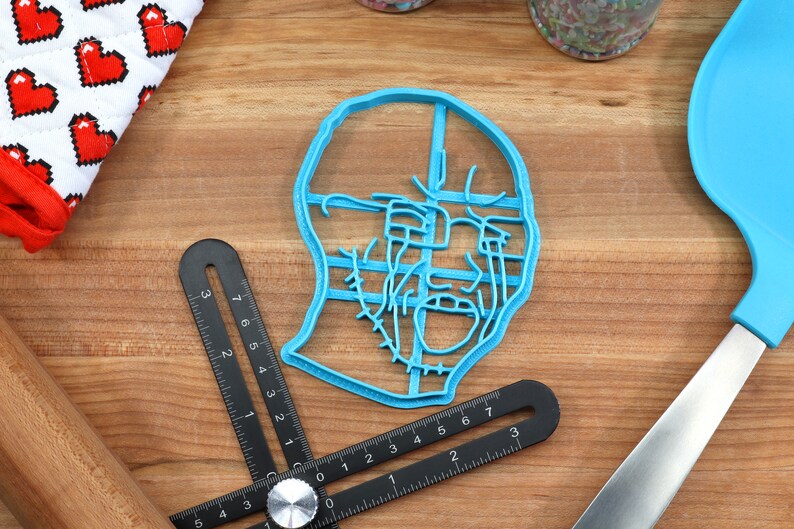 May include: A blue plastic cookie cutter shaped like a face with a detailed design. The cutter is on a wooden surface with a ruler and a spatula.