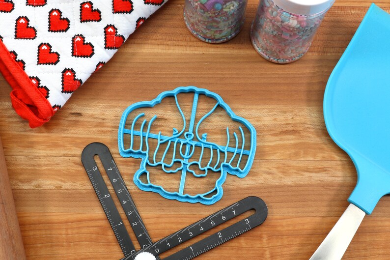 May include: Blue plastic cookie cutter shaped like a cartoon dog wearing a sweater. The cutter is on a wooden surface.