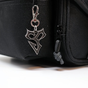 May include: A black zippered bag with a silver keychain featuring a stylized symbol. The bag has a gray strap and a black buckle.