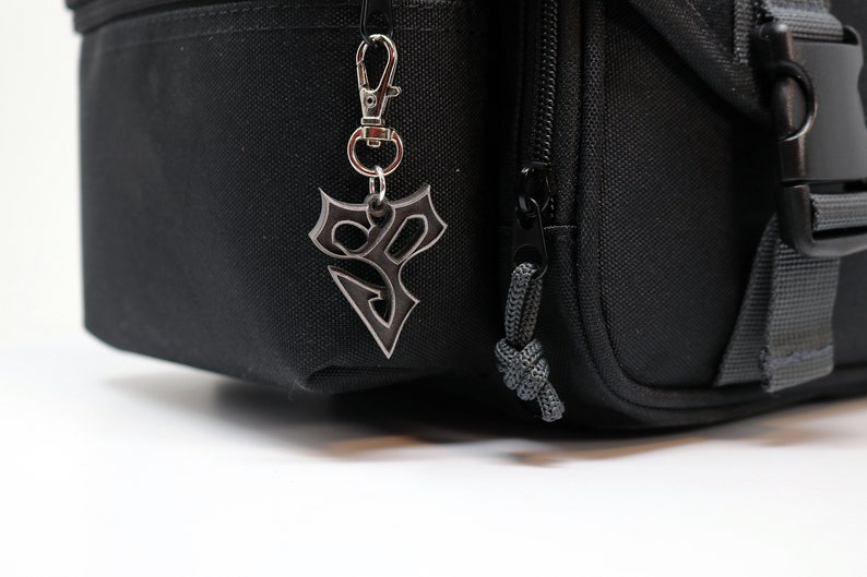 May include: A black zippered bag with a silver keychain featuring a stylized symbol. The bag has a gray strap and a black buckle.