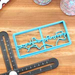 May include: A blue plastic cookie cutter shaped like a city skyline. The cutter is on a wooden surface.