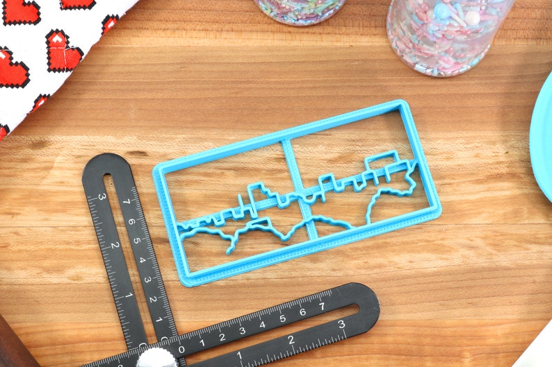 May include: A blue plastic cookie cutter shaped like a city skyline. The cutter is on a wooden surface.
