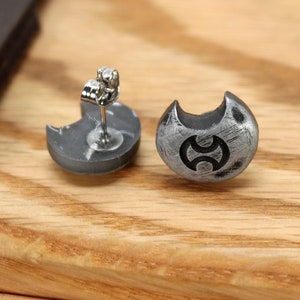 May include: A pair of silver earrings with a crescent moon design and a stylised symbol in the centre. The earrings are on a wooden surface.