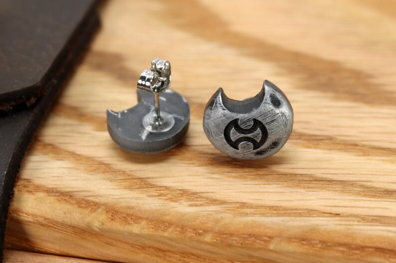 May include: A pair of silver earrings with a crescent moon design and a stylised symbol in the centre. The earrings are on a wooden surface.
