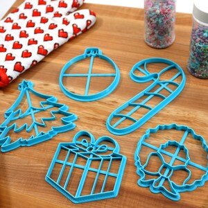 Op de afbeelding: Een set van zes blauwe plastic kerstkoekjesvormen in de vorm van een kerstboom, een cadeau, een snoepstok, een krans, een ornament en een ster. De vormen liggen op een houten oppervlak met sprinkles in potten en een deegroller.