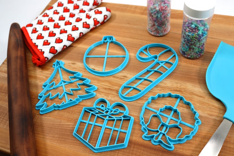 Op de afbeelding: Een set van zes blauwe plastic kerstkoekjesvormen in de vorm van een kerstboom, een cadeau, een snoepstok, een krans, een ornament en een ster. De vormen liggen op een houten oppervlak met sprinkles in potten en een deegroller.