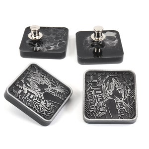 Puede incluir: Cuatro botones cuadrados negros con un dise&ntilde;o plateado. Dos botones tienen un pasador plateado en la parte superior. Los otros dos botones tienen un dise&ntilde;o plateado de una mujer con texto japon&eacute;s.