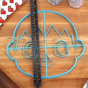 May include: A blue plastic cookie cutter shaped like a cartoon face with a smile, eyes, and hair. The cutter is on a wooden surface with a ruler and a spatula.