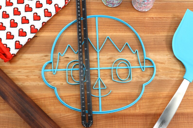 May include: A blue plastic cookie cutter shaped like a cartoon face with a smile, eyes, and hair. The cutter is on a wooden surface with a ruler and a spatula.