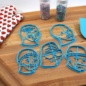 May include: A set of five blue plastic cookie cutters shaped like different faces. The cutters are on a wooden cutting board with a rolling pin, a spatula, and two jars of sprinkles.