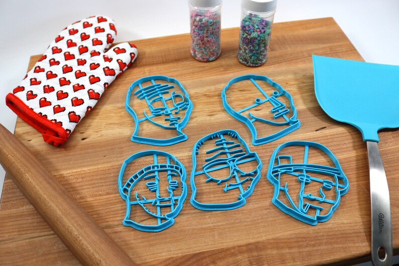 May include: A set of five blue plastic cookie cutters shaped like different faces. The cutters are on a wooden cutting board with a rolling pin, a spatula, and two jars of sprinkles.