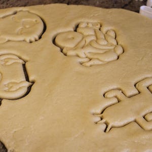 May include: Close-up of cookie dough with several animal-shaped cutouts. The cutouts include a frog, a pig, and other animal shapes. A white plastic cookie cutter is visible in the background.