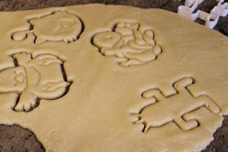 May include: Close-up of cookie dough with several animal-shaped cutouts. The cutouts include a frog, a pig, and other animal shapes. A white plastic cookie cutter is visible in the background.