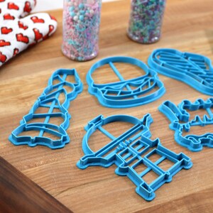 K&ouml;nnte beinhalten: F&uuml;nf blaue Plastik-Ausstecher in Form eines Hamburgers, eines Leuchtturms, eines Grills, einer Geburtstagstorte und eines Weihnachtsbaums. Die Ausstecher liegen auf einer Holzoberfl&auml;che.