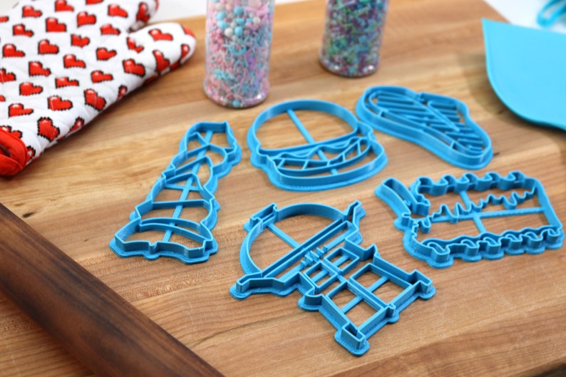 K&ouml;nnte beinhalten: F&uuml;nf blaue Plastik-Ausstecher in Form eines Hamburgers, eines Leuchtturms, eines Grills, einer Geburtstagstorte und eines Weihnachtsbaums. Die Ausstecher liegen auf einer Holzoberfl&auml;che.