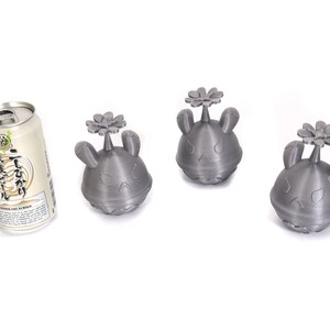 May include: Three gray 3D printed figurines with a flower on top. The figurines have a cartoonish face with a frown. A can of Japanese beer is in the background.