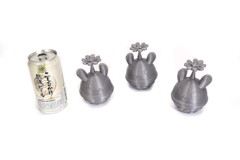 May include: Three gray 3D printed figurines with a flower on top. The figurines have a cartoonish face with a frown. A can of Japanese beer is in the background.