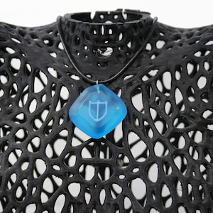 Puede incluir: Un collar con un colgante de cristal azul con un dise&ntilde;o de escudo blanco. El collar est&aacute; en un maniqu&iacute; negro.