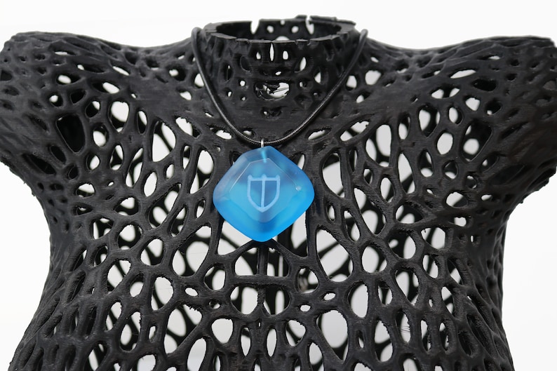 Puede incluir: Un collar con un colgante de cristal azul con un dise&ntilde;o de escudo blanco. El collar est&aacute; en un maniqu&iacute; negro.