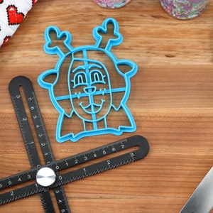 Peut inclure: Un emporte-pi&egrave;ce en plastique bleu en forme de renne avec un visage souriant et des bois. Un outil de mesure multi-angles noir est positionn&eacute; pr&egrave;s du cutter. Un gant de cuisine &agrave; motifs de c&oelig;urs rouges et blancs est visible.